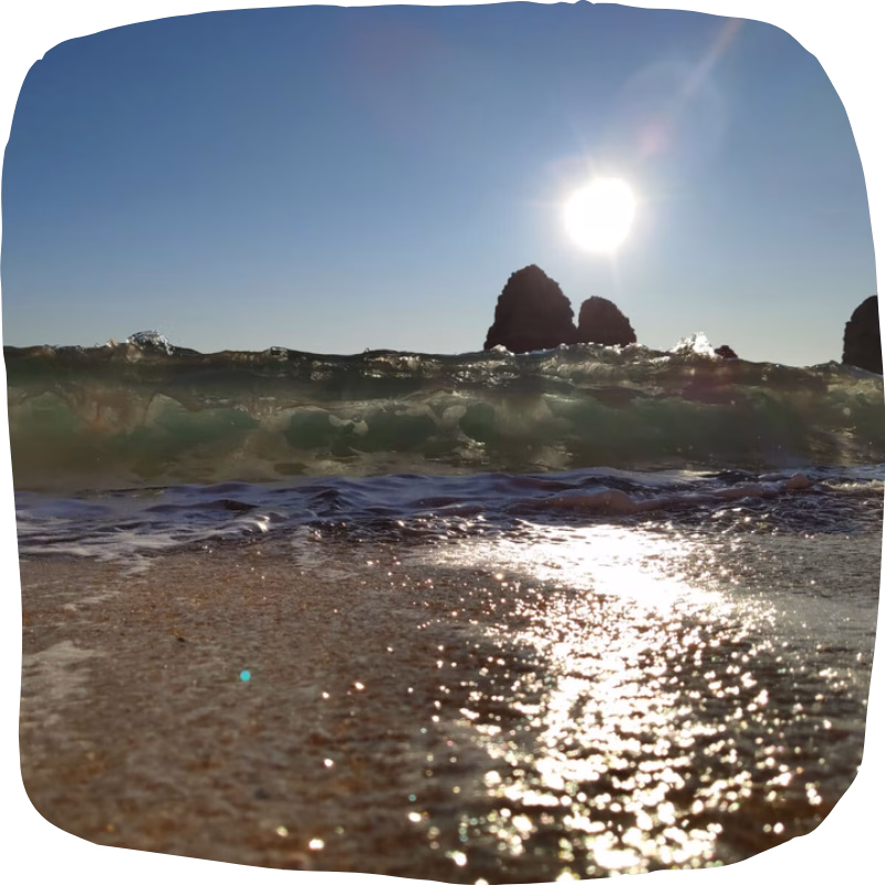 Low-angle view from the sand of an Algarve beach with a breaking wave, the sun shining above and rocky formations in the background